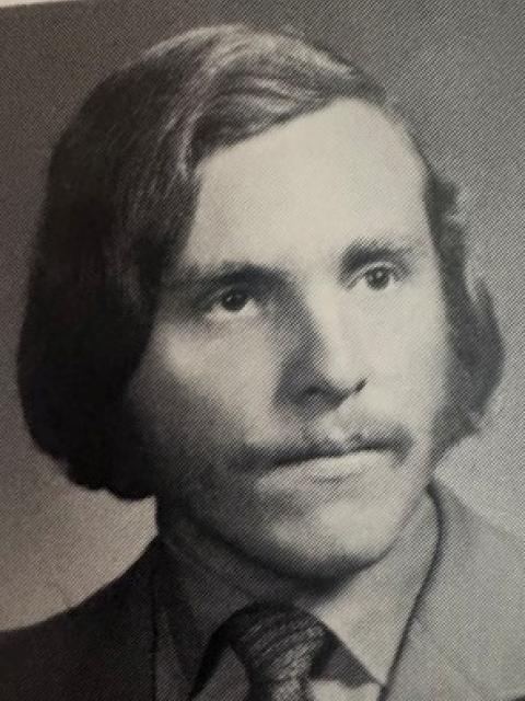 Black-and-white yearbook portrait of a person wearing a suit jacket, collared shirt and patterned tie. The person is shown from the shoulders up, facing slightly to the side.