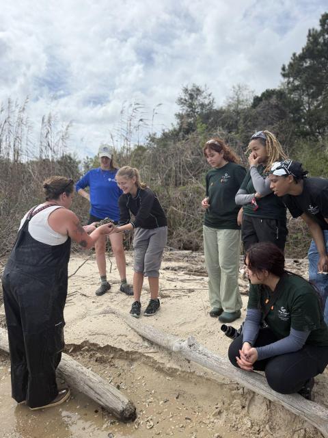 Small group of students and instructors gather on a sandy shoreline to examine oyster samples during a field experience.