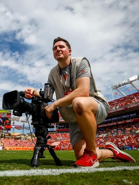 A camera operator kneels on the sideline of a football field beside a professional video camera before a game.