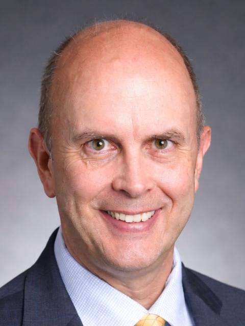 Professional headshot of Eric Johnson wearing a dark suit jacket, light dress shirt and patterned tie against a neutral gray background.