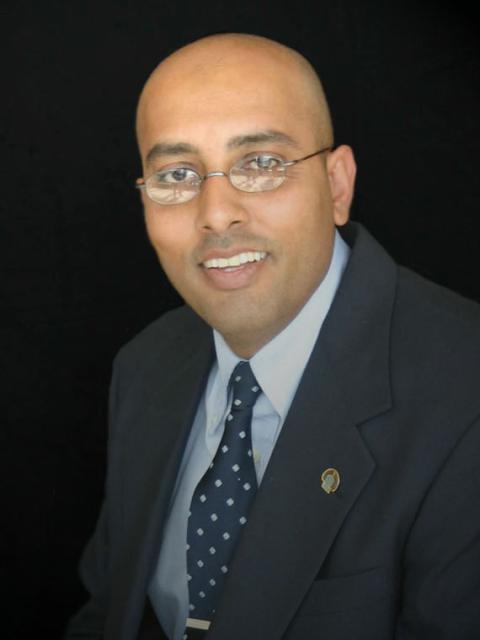 Formal portrait of a person wearing a dark suit jacket, light blue shirt and navy patterned tie, seated against a black background and looking toward the camera.